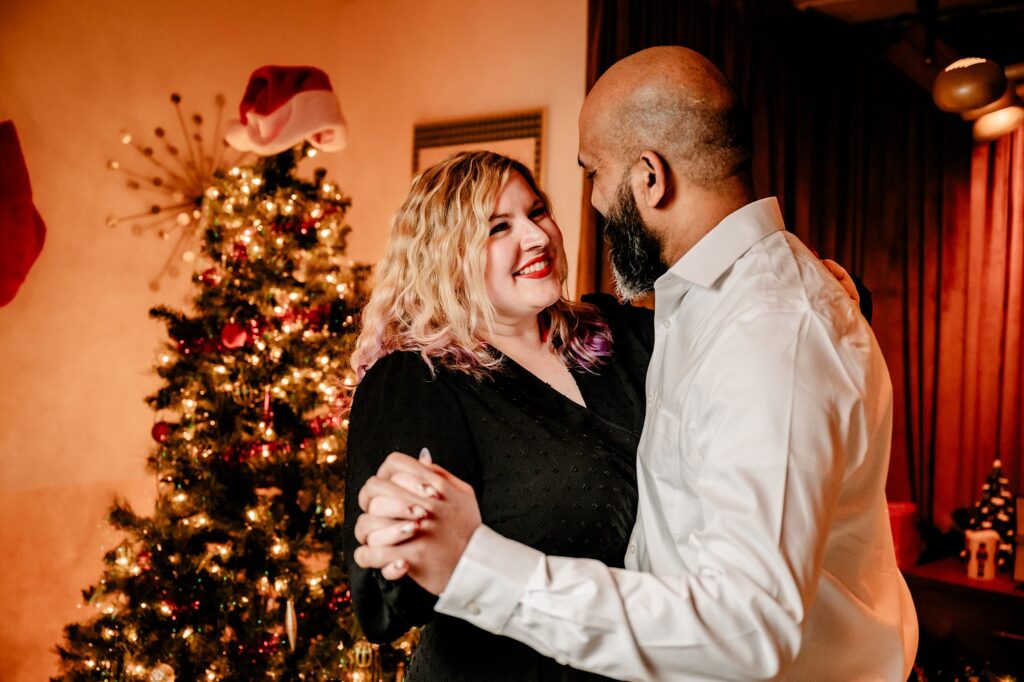 A smiling couple dances together in front of a decorated Christmas tree with lights and ornaments, capturing classic Christmas vibes. The woman wears a black dress and the man a white shirt, creating a warm and festive holiday couples portrait.