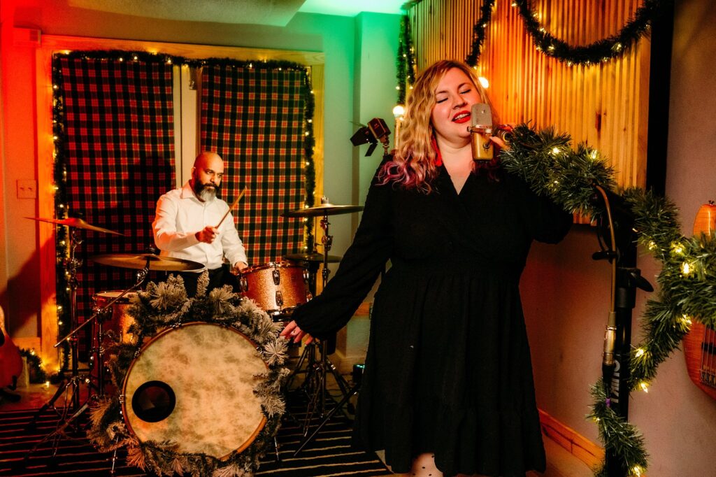 A woman in a black dress sings into a gold microphone decorated with garland, while a drummer plays behind her. The festively lit room with string lights and plaid curtains sets the scene for a cozy, Vintage Christmas vibe.