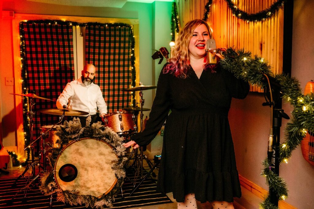 A woman in a black dress sings into a garland-wrapped microphone, while a man plays drums behind her. The warmly lit room, adorned with string lights and festive touches, evokes the charm of a Vintage Christmas.