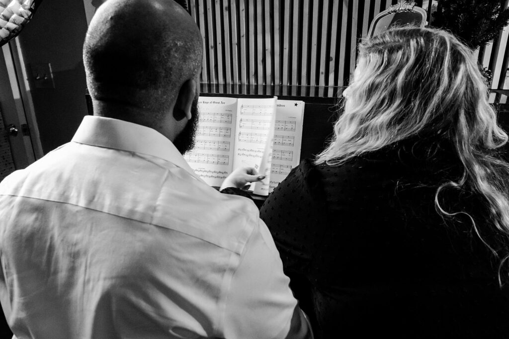 Two people, seen from behind, sit side by side looking at sheet music on a stand—one points to the music with a finger. This black and white image evokes a cozy, vintage Christmas feel—perfect for a holiday couples portrait.