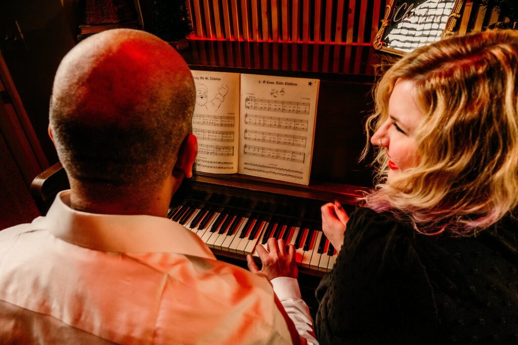 A man and a woman sit at a piano, viewed from behind. The man plays while the blonde woman with red lipstick smiles at him. Sheet music is open on the piano in this cozy Holiday Couples Portrait.