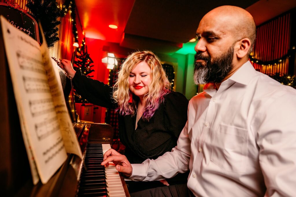 A woman smiles while turning sheet music on a piano as a man in a white shirt plays; colorful holiday lights and festive decorations create the perfect setting for a Vintage Christmas couples portrait.