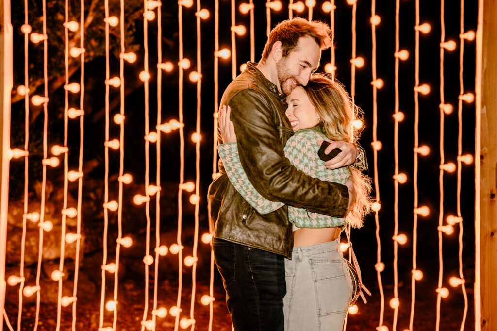 A couple embraces and smiles in front of a wall of hanging string lights at night, creating a warm and romantic atmosphere—perfect for a Surprise Proposal captured by a Nashville Proposal Photographer.