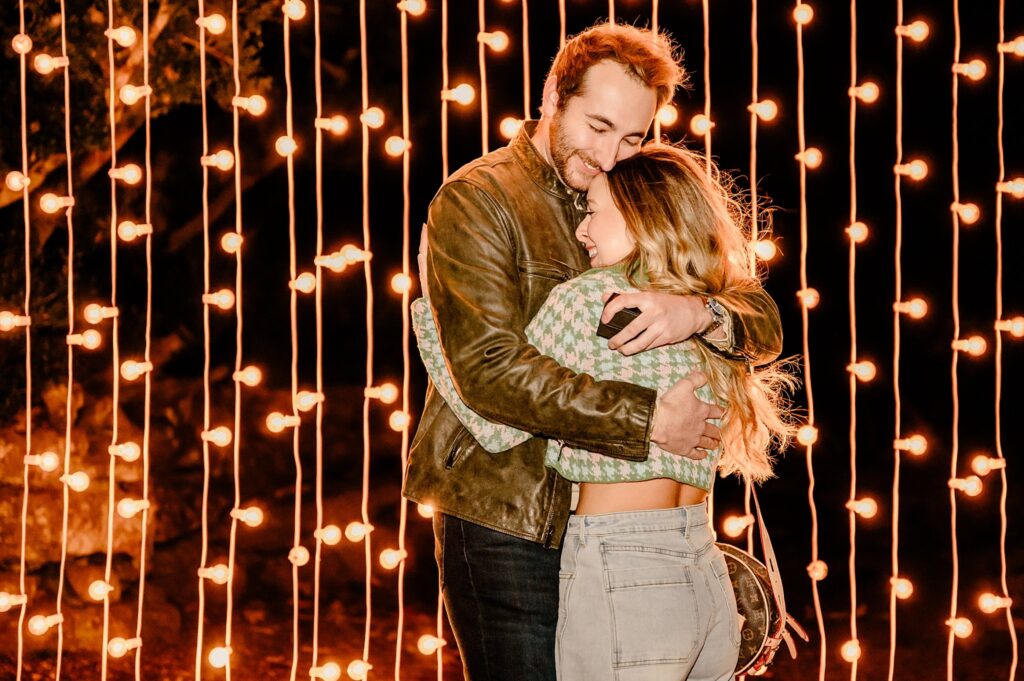 A couple hugs and smiles warmly in front of a curtain of glowing string lights at night, creating a cozy and romantic atmosphere—perfect for a surprise proposal captured by a Nashville Proposal Photographer.