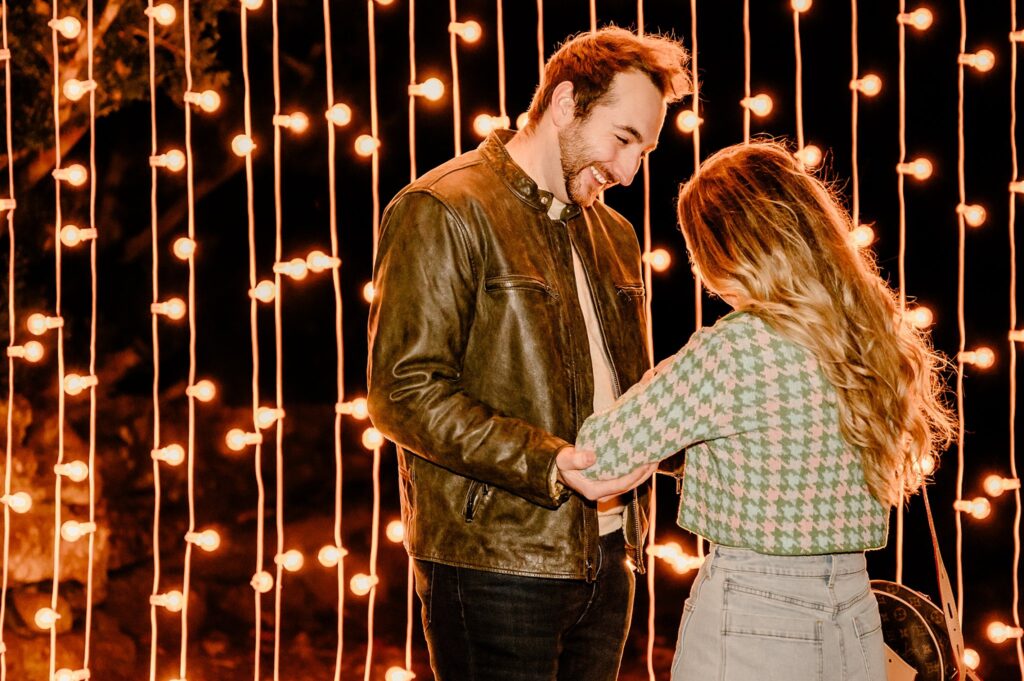 A man and woman stand in front of glowing string lights at night, smiling and holding hands. The man wears a brown jacket, and the woman, in a green patterned sweater, beams—captured by a Nashville Proposal Photographer during their Surprise Proposal.