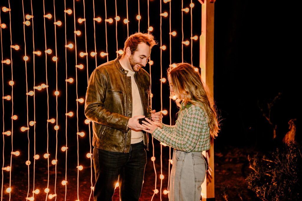 A man and woman stand in front of glowing string lights at night. The man, smiling, holds a small open box while the woman looks down happily—it’s a magical surprise proposal captured perfectly by a Nashville Proposal Photographer at Cheekwood.