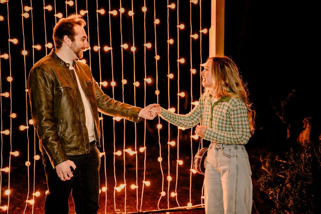 A man and woman stand in front of hanging string lights at night, holding hands and smiling at each other. The scene is warmly lit, giving a festive and intimate atmosphere—perfect for a Cheekwood surprise proposal.