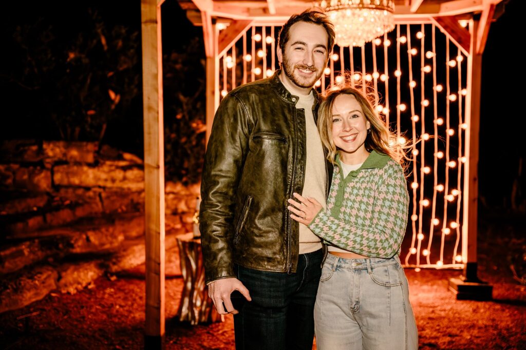 A man and woman stand smiling together under a gazebo decorated with hanging string lights at night. Captured by a Nashville Proposal Photographer, the moment feels magical as they celebrate in style—he in a leather jacket, she in green and jeans.