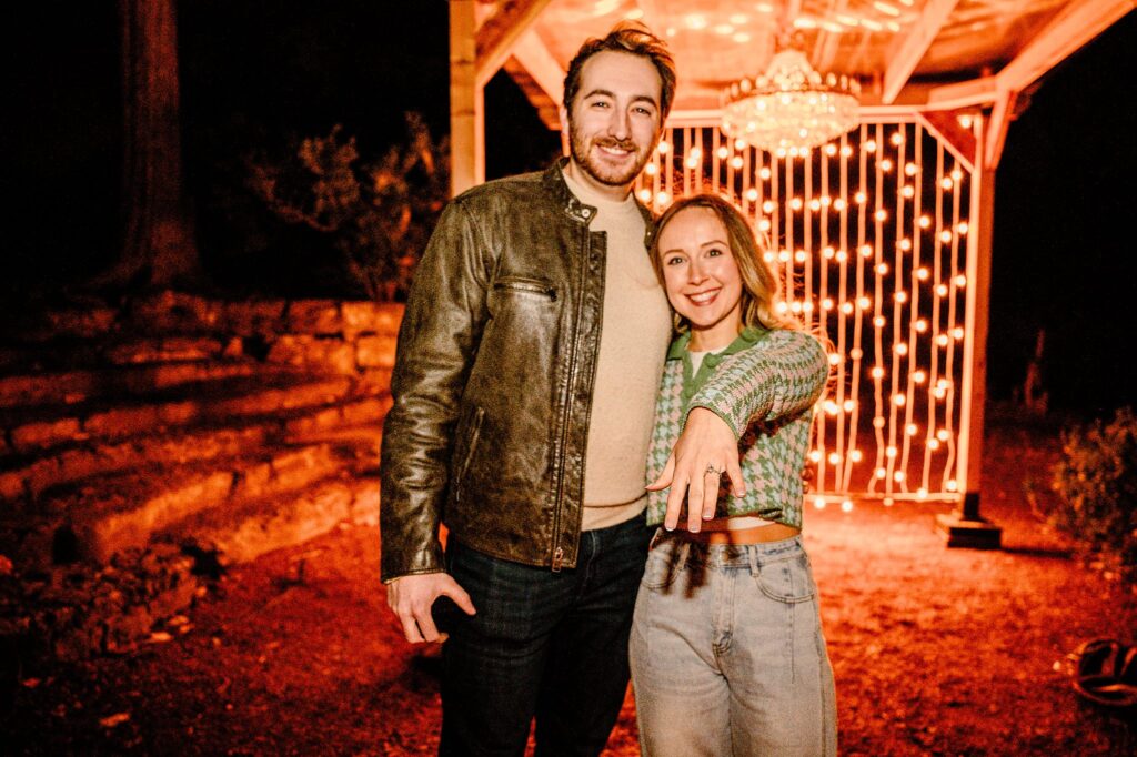A smiling couple poses outside at night under string lights. The woman extends her hand forward, showing a ring, while the man stands beside her with his arm around her shoulders—captured by a Nashville Proposal Photographer.