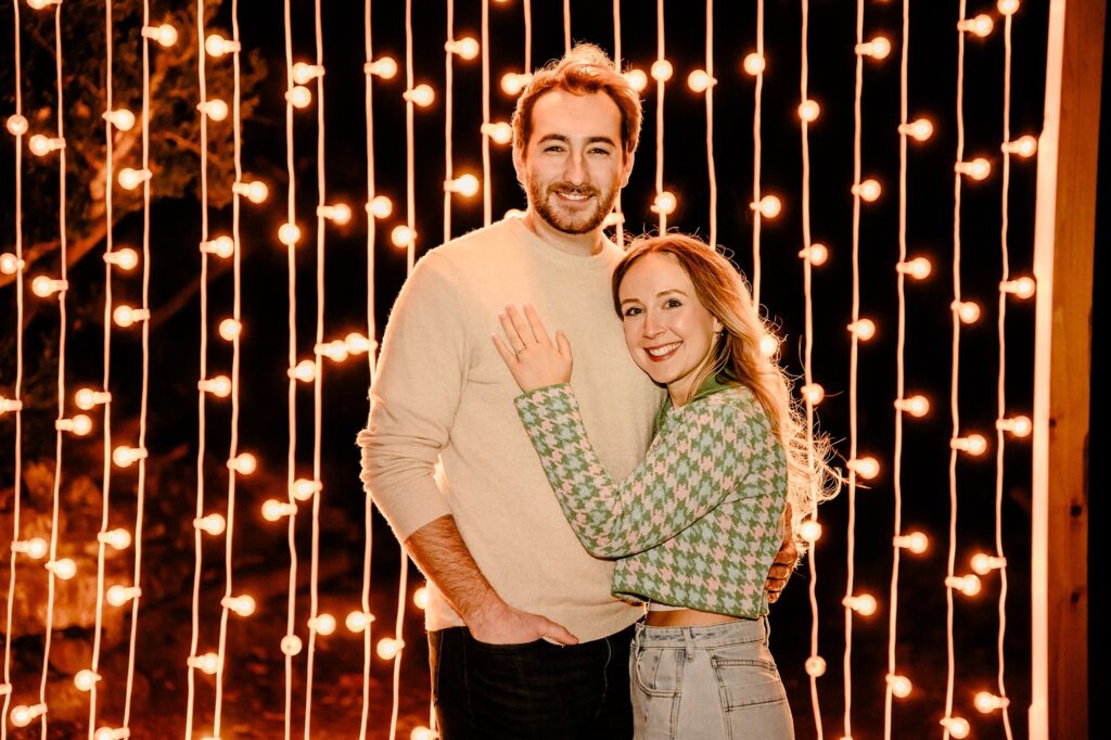 A smiling couple stands together at night in front of vertical rows of glowing string lights at Cheekwood. The woman embraces the man’s waist as they both look at the camera, creating a warm, festive atmosphere for their surprise proposal.