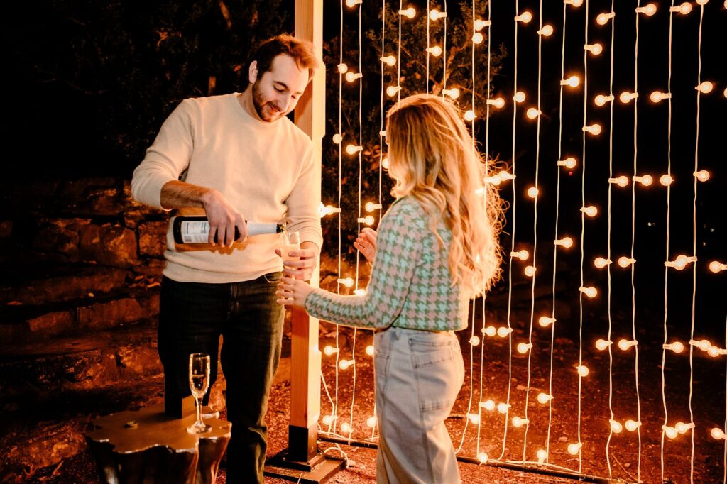 A man pours wine into a glass held by a woman as they stand outdoors at night, illuminated by string lights. This warm, festive moment could be perfectly captured by a Nashville Proposal Photographer at Cheekwood.