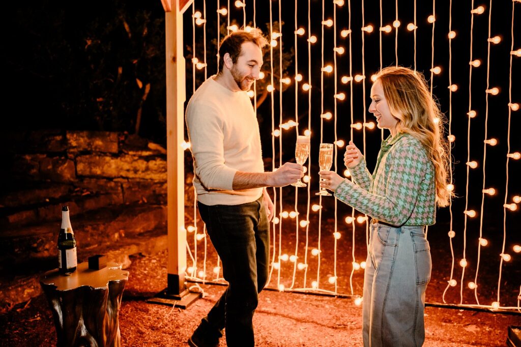 A man and woman stand smiling and clinking champagne glasses outdoors at night, a string of warm lights behind them, celebrating a surprise proposal. Captured by a Nashville Proposal Photographer at Cheekwood, a bottle sits on the table nearby.