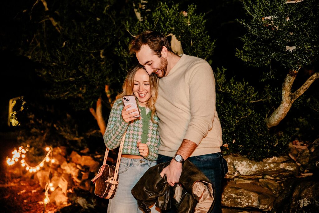 A smiling couple stands close together outdoors at night, looking at a phone in the woman’s hand. String lights and greenery create a warm, festive atmosphere—perfect for a surprise proposal captured by a Nashville Proposal Photographer at Cheekwood.