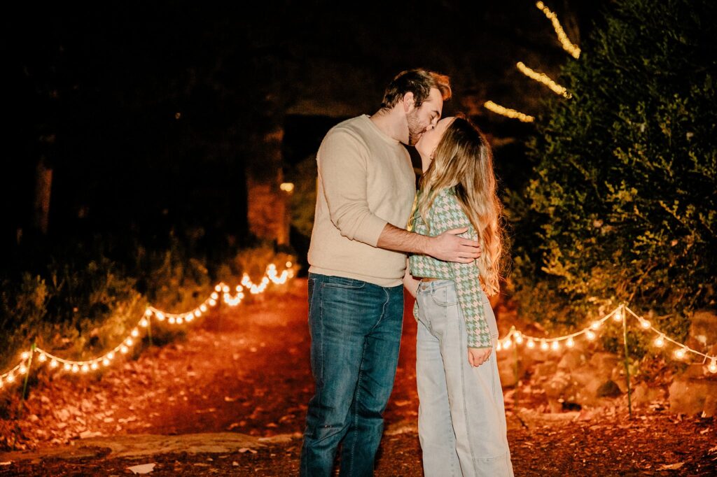 A couple stands on a leaf-covered path at Cheekwood, sharing a kiss under string lights. The man wears jeans and a beige sweater; the woman, a green plaid shirt and light blue jeans—a magical moment captured by a Nashville Proposal Photographer.