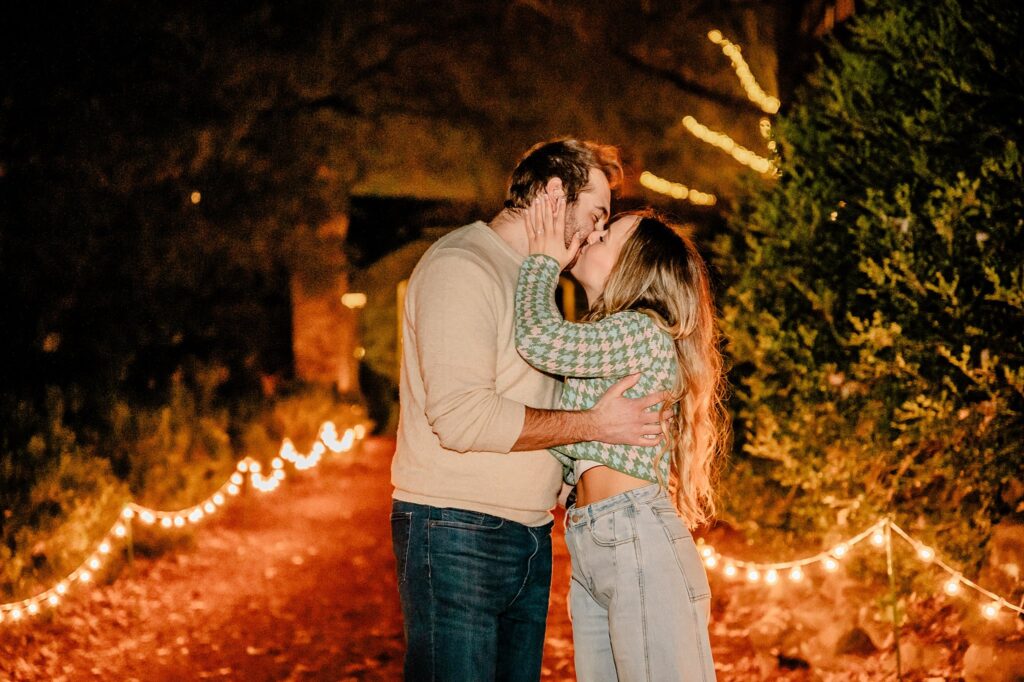 A couple stands on a path lined with string lights at night, embracing and kissing beneath the trees. The scene is warmly lit, creating a romantic atmosphere—perfect for a Cheekwood surprise proposal captured by a Nashville Proposal Photographer.