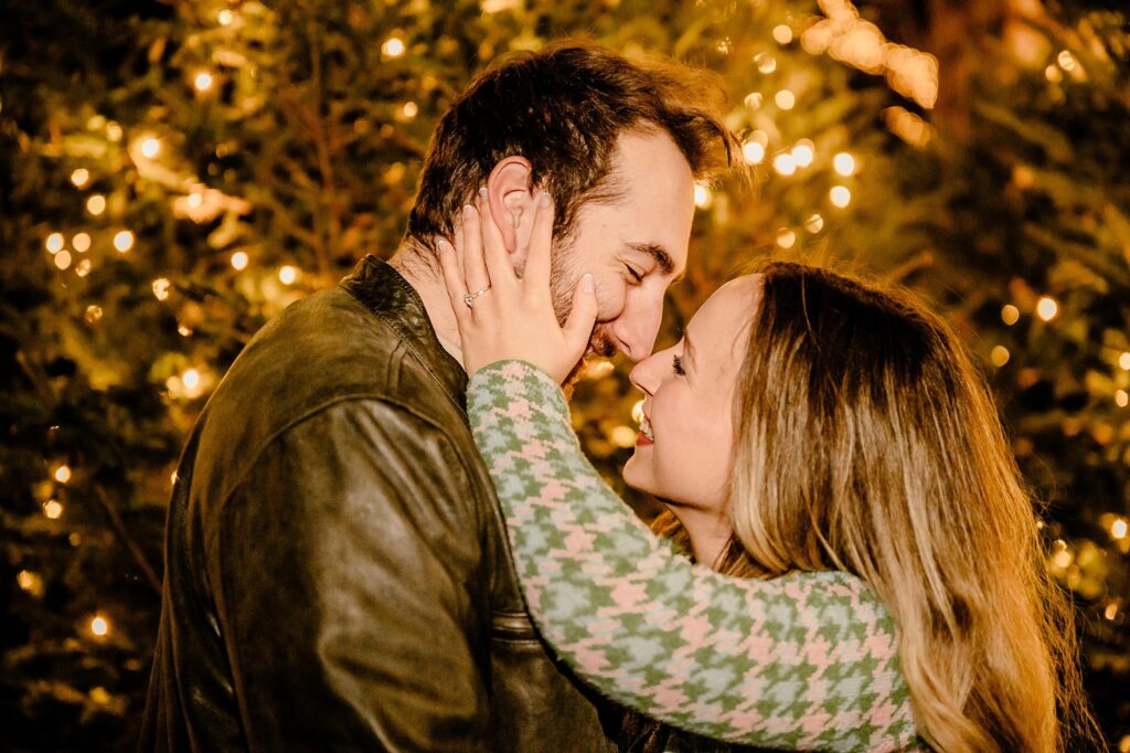 A couple smiles closely at each other, standing in front of twinkling lights and evergreen trees at Cheekwood. The woman gently holds the man's face, both looking happy and affectionate during their magical surprise proposal.