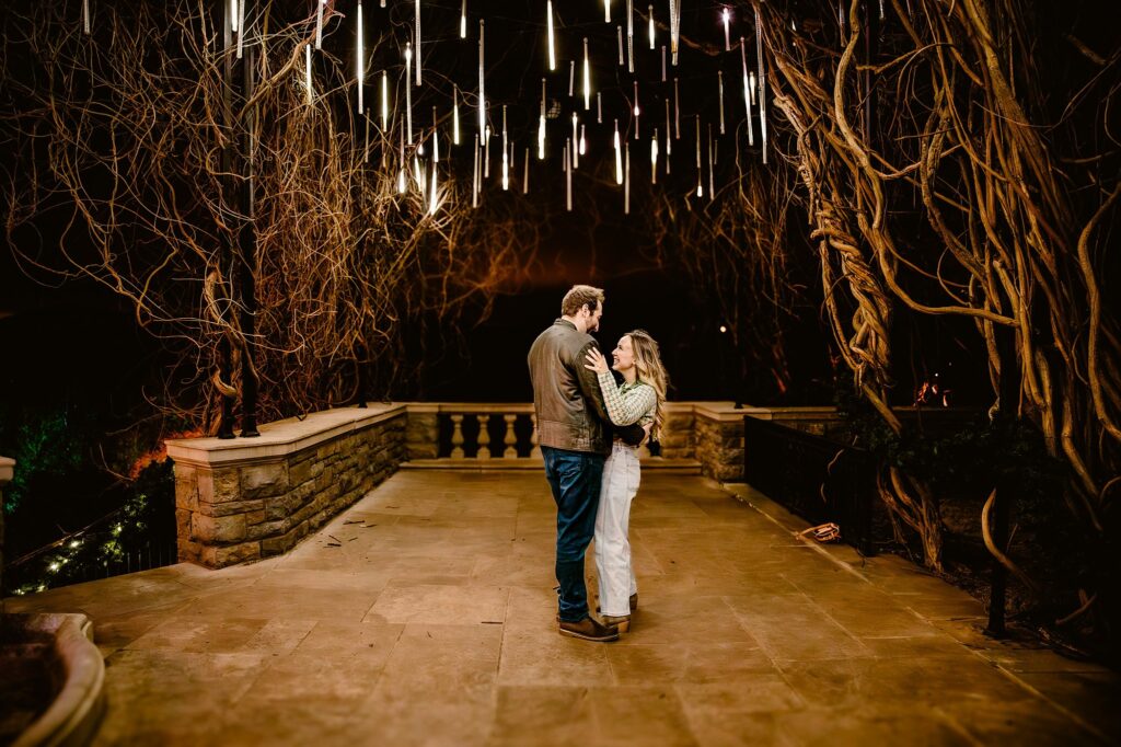 A couple embraces on a stone terrace at night at Cheekwood, surrounded by leafless twisted vines and glowing hanging lights—an enchanting scene perfect for a surprise proposal, beautifully captured by a Nashville Proposal Photographer.