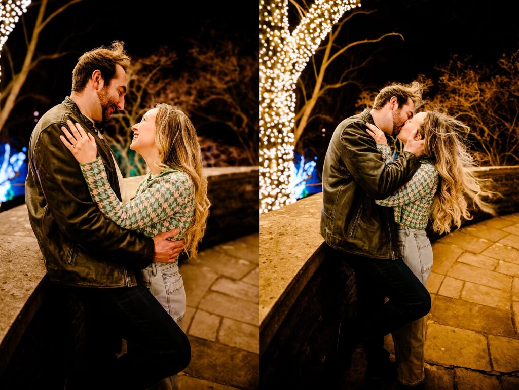 A couple embraces and kisses on a stone walkway at night at Cheekwood, surrounded by trees wrapped in glowing string lights. They look happy and affectionate, sharing a romantic outdoor moment captured by a Nashville proposal photographer.