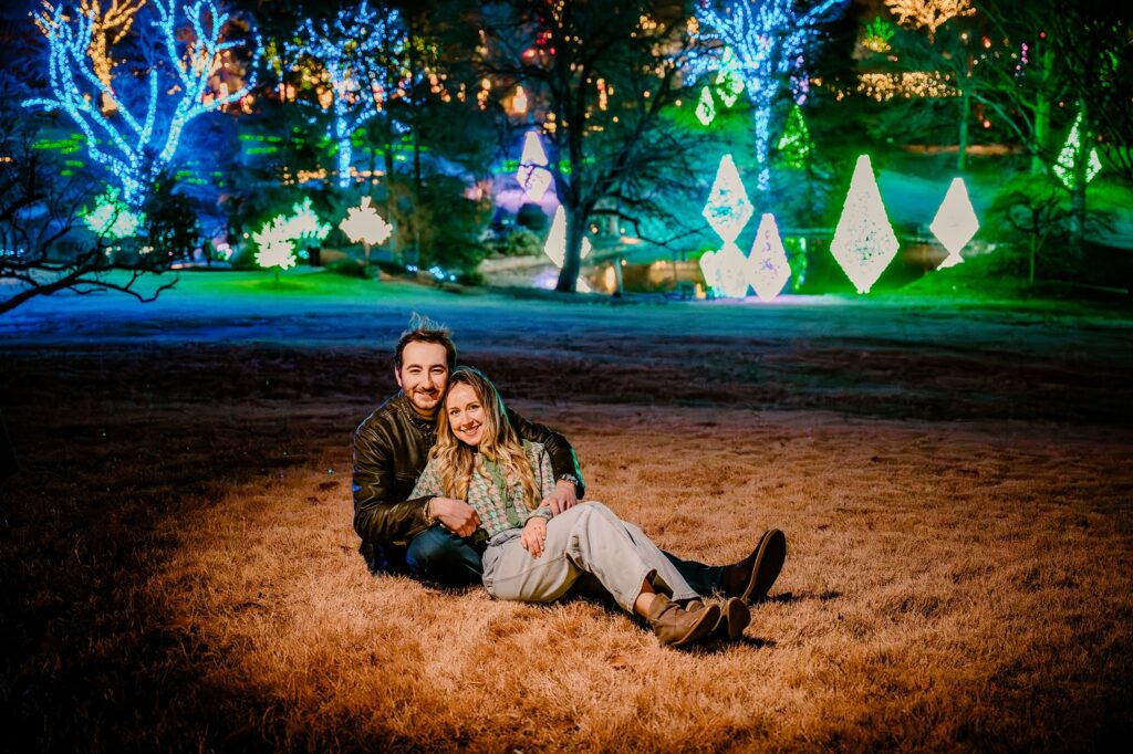 A couple sits on grass at night, smiling and leaning against each other at Cheekwood, surrounded by colorful, glowing holiday lights and illuminated decorations in the background.