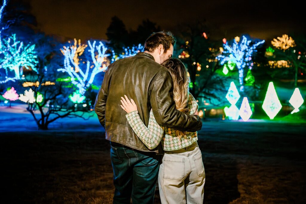 A couple stands close together outdoors at night at Cheekwood, arms around each other, looking at brightly lit trees and glowing decorations in a festive, illuminated park—perfect for a Nashville Proposal Photographer to capture a magical surprise proposal.