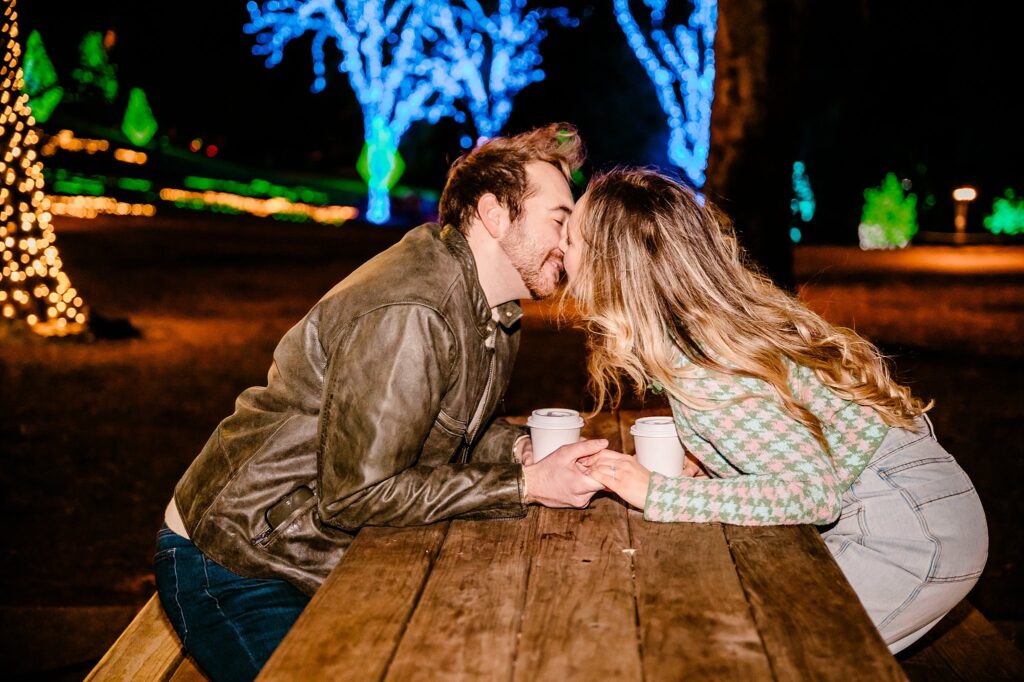A couple sits at a wooden picnic table at night, holding hands and leaning in for a kiss with coffee cups, as colorful holiday lights illuminate the trees—perfectly capturing a Surprise Proposal by a Nashville Proposal Photographer.