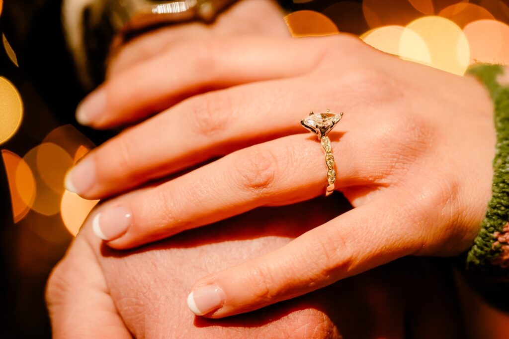 A close-up of a hand with a diamond engagement ring, gently resting on another's, captures the magic of a surprise proposal, with warm, blurred lights in the background—perfectly documented by a Nashville Proposal Photographer.