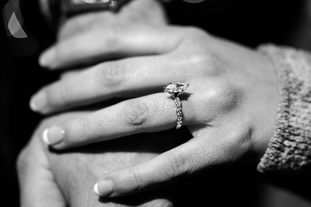 Close-up of a hand wearing an engagement ring with a diamond on the ring finger, gently resting on another's hand. Captured in black and white by a Nashville Proposal Photographer, this image beautifully celebrates a surprise proposal.