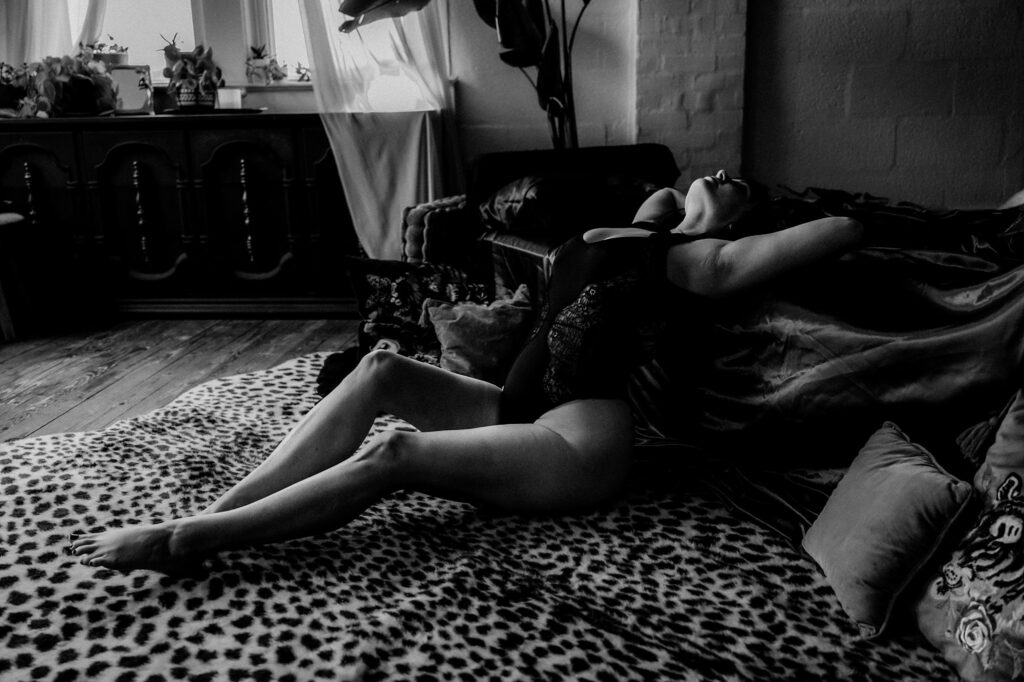 A woman in a black bodysuit reclines on a sofa draped with a leopard-print blanket, arching her back in a dimly lit, stylish room. This striking black and white scene captures the essence of Nashville Boudoir Photography.