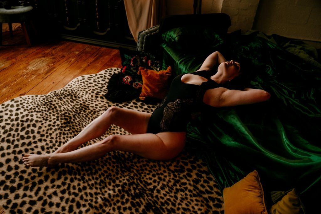 A woman in a black lace bodysuit reclines on a leopard-print rug by a green velvet bed, her arms raised above her head. Warm wood floors and decorative pillows complete this moody Nashville Boudoir Photography scene.