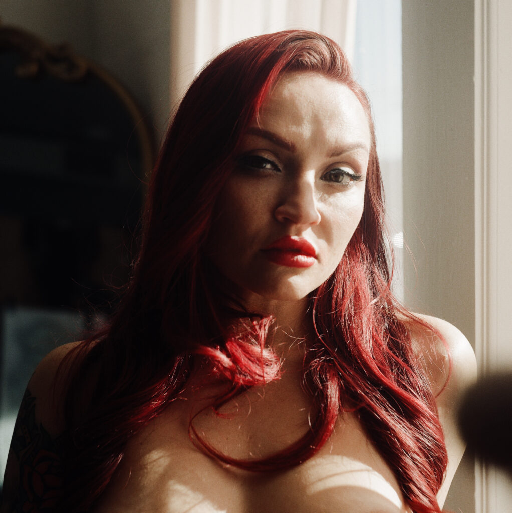 A woman with long red hair and red lipstick stands by a sunlit window, looking directly at the camera with a serious expression. This Nashville boudoir moment captures light softly highlighting her hair and face.