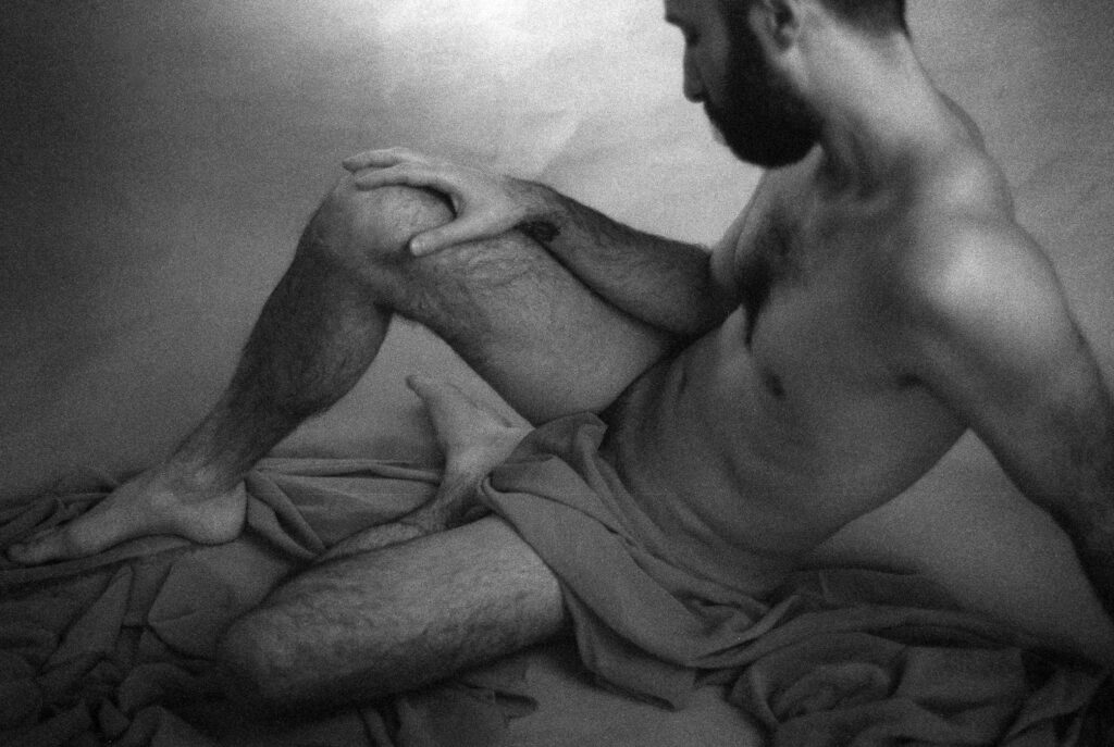 Black and white photo of a bearded man sitting on draped fabric, partially nude, his knee raised and arm resting on his leg, gazing downward. This Nashville boudoir image emphasizes form and shadow.