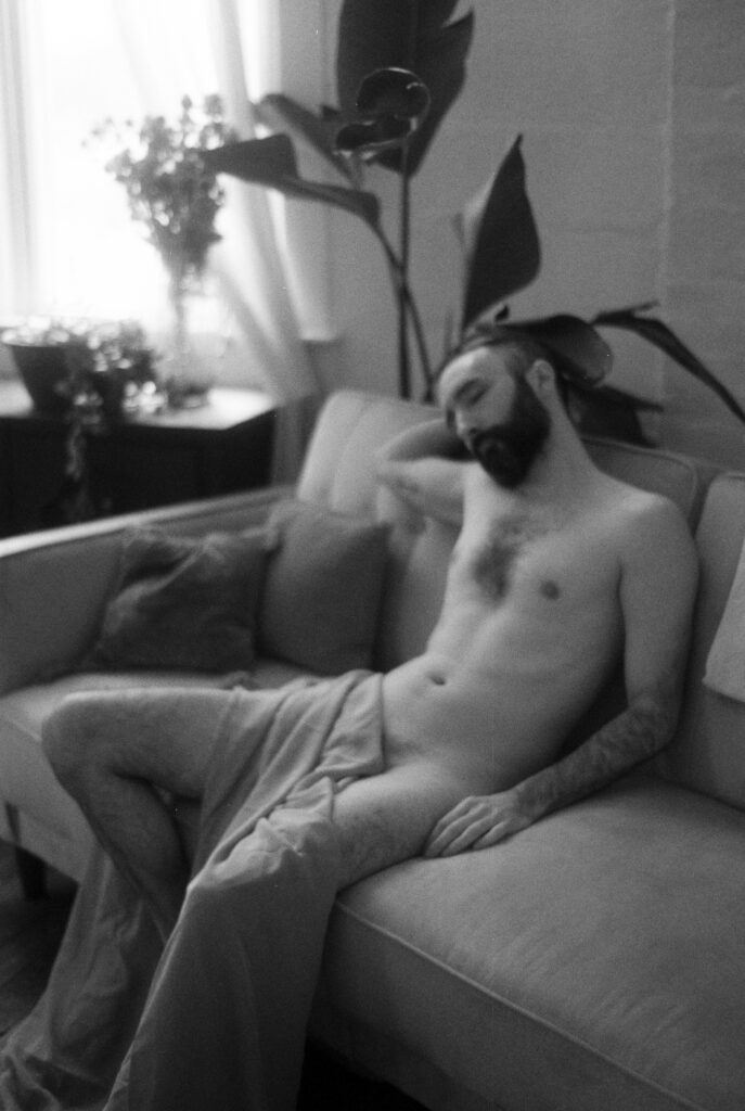 A black-and-white photo of a shirtless person with a beard sitting on a couch, partially covered by a blanket in this Nashville boudoir scene. Sunlight filters through the window, highlighting houseplants in the background.