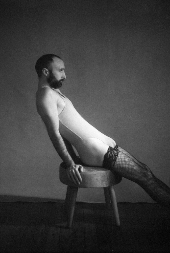 A person with a beard and short hair, wearing a bodysuit, fishnet stockings, and lace garters, leans back on a round stool in a dimly lit room with a plain background, capturing edgy nashville boudoir vibes.
