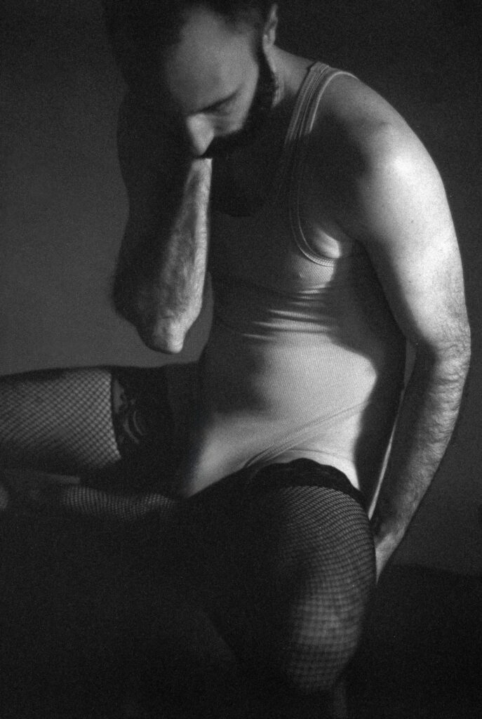 A person with a beard sits thoughtfully, wearing a tank top and fishnet stockings. This Nashville boudoir black and white photo uses dramatic lighting and shadow to create a contemplative, moody atmosphere.