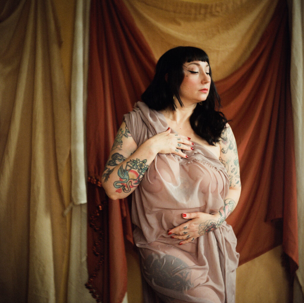 A person with black hair and visible tattoos poses with eyes closed, draped in sheer, taupe fabric. Captured in a Nashville boudoir style, they stand before layered, earth-toned cloth backdrops for an intimate, artistic vibe.