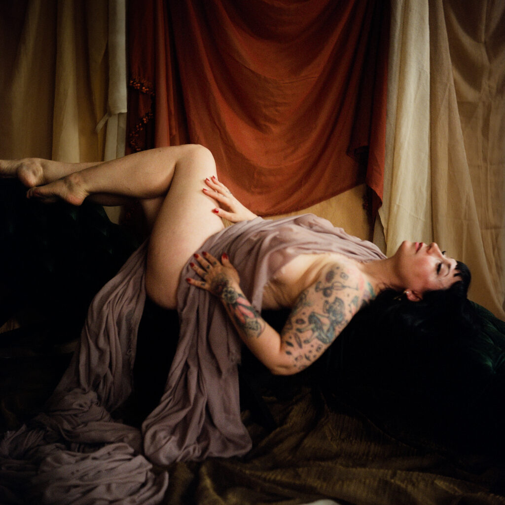 A person with tattoos reclines on a dark surface, draped in sheer, light fabric. With dark hair and red nails, they are bathed in soft lighting—capturing the intimate elegance of a Nashville boudoir among earthy-toned curtains.