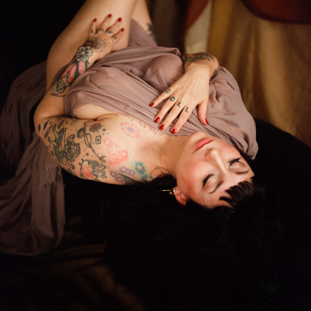 A woman with tattoos and red nail polish lies on her back with her eyes closed, draped in a soft, sheer fabric—an intimate Nashville boudoir scene—one hand on her chest and the other near her face in gentle, dreamy light.
