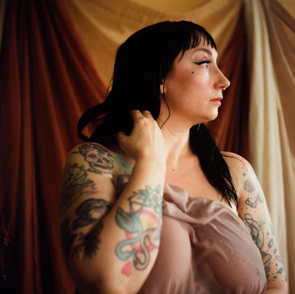 A woman with long dark hair and visible tattoos on her arms stands in front of draped, earth-toned fabric. She gazes to the side and gently touches her hair, embodying a soft, sheer look for this Nashville boudoir session.