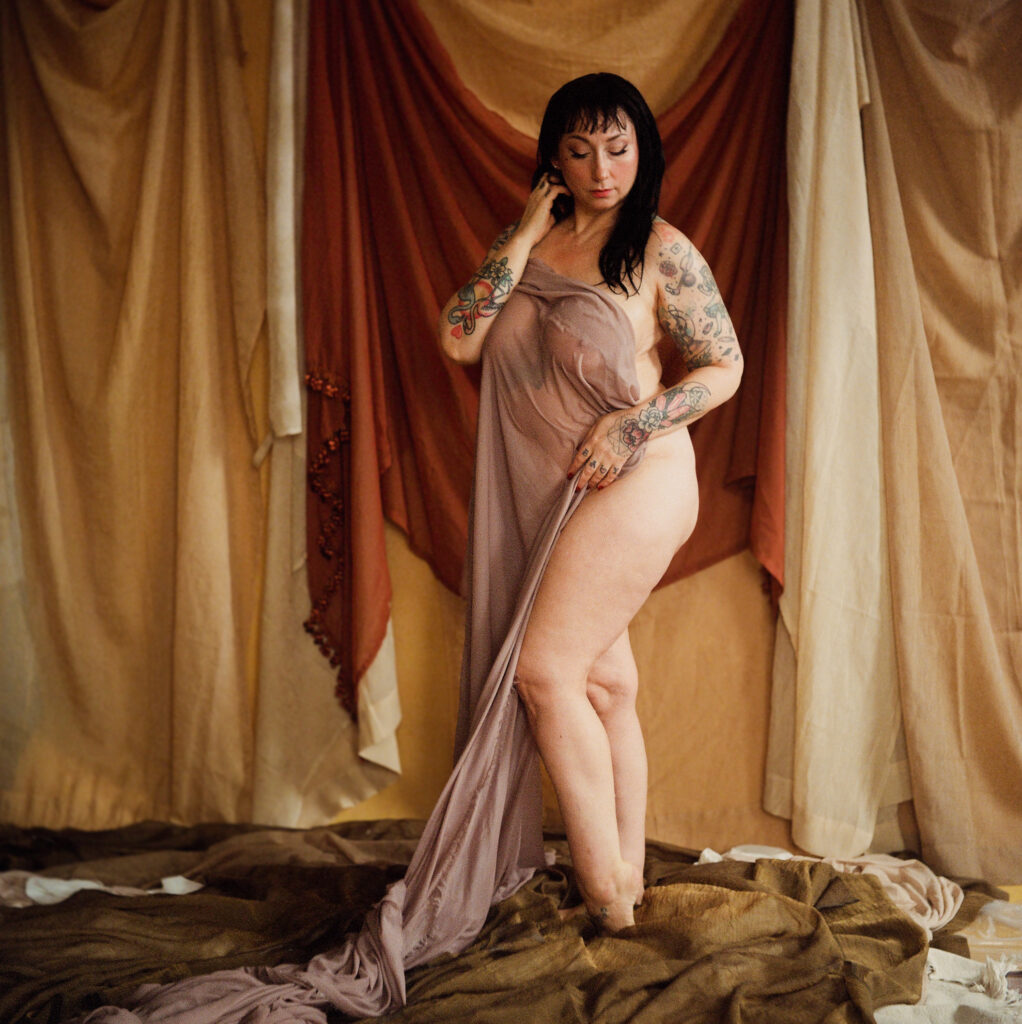 A person with tattoos drapes a sheer, mauve fabric around themselves, standing barefoot on cloth, set against muted brown and beige fabrics—a dreamy Nashville boudoir scene.