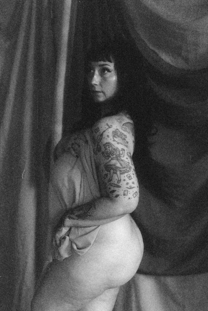 Black and white photo of a person with long dark hair and visible tattoos on their arm, posing with fabric draped around their body against a softly draped backdrop, capturing the intimate style of Nashville boudoir photography.