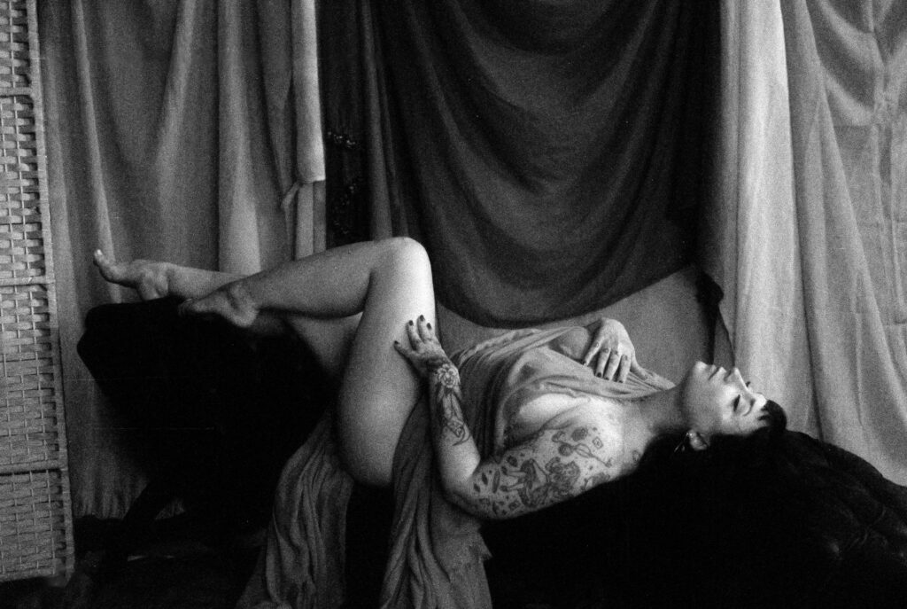 A person with tattoos reclines on a chair, draped in fabric, looking up with eyes closed. The Nashville boudoir setting features soft, flowing curtains and a wicker screen in black and white, creating a dramatic, artistic mood.
