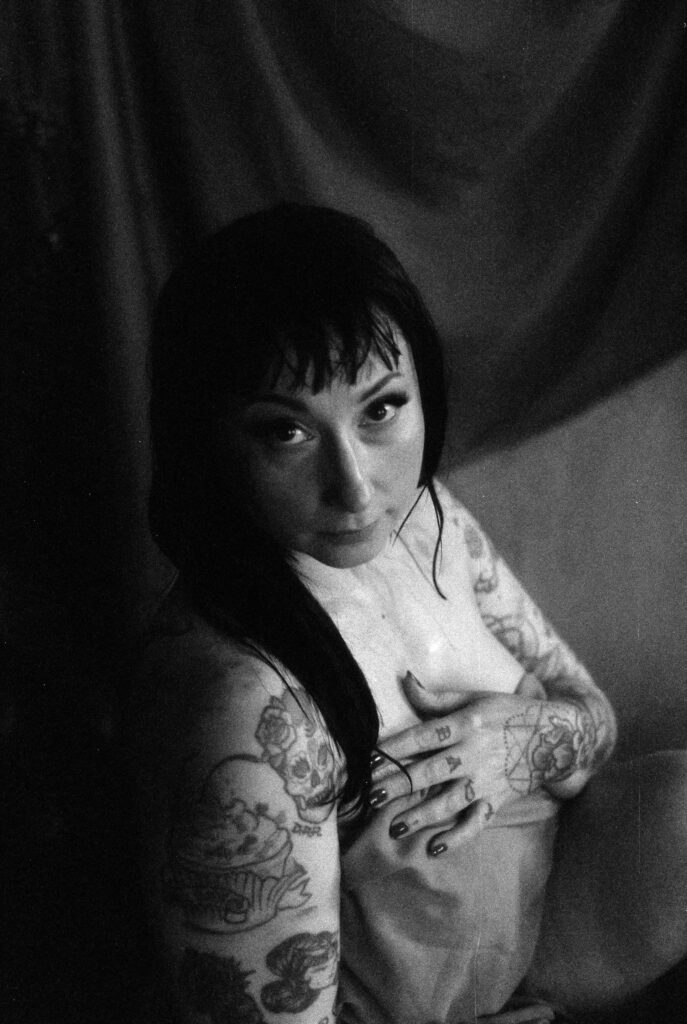 A black and white nashville boudoir photo of a person with wet hair, tattoos on their arms and hands, and dark eye makeup, sitting against draped fabric while looking up at the camera and covering their chest with their hand.