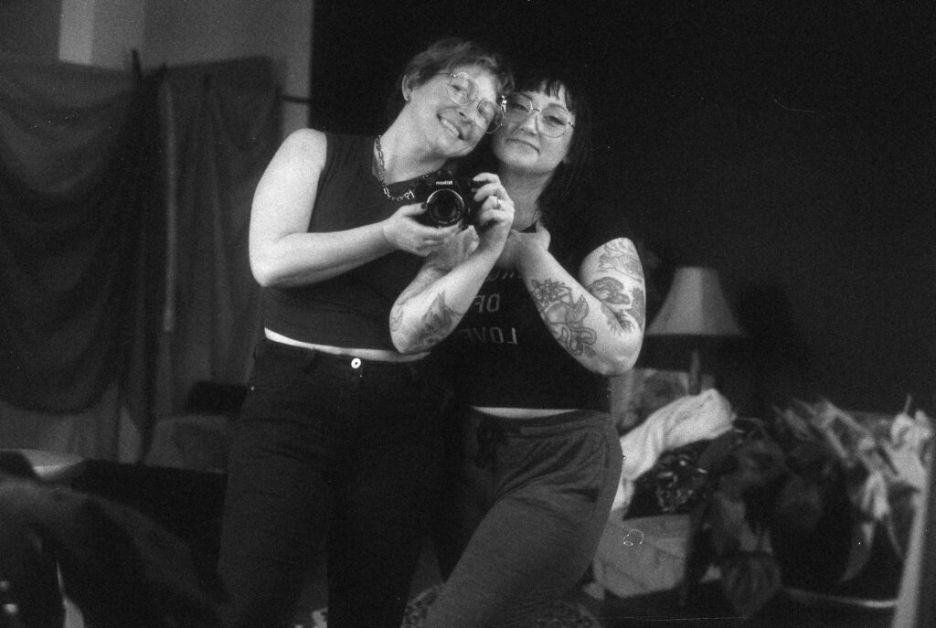 Two people with glasses, tattoos, and short hair smile while posing together for a mirror selfie in a cozy room with a lamp and piles of clothes—a casual Nashville boudoir moment.