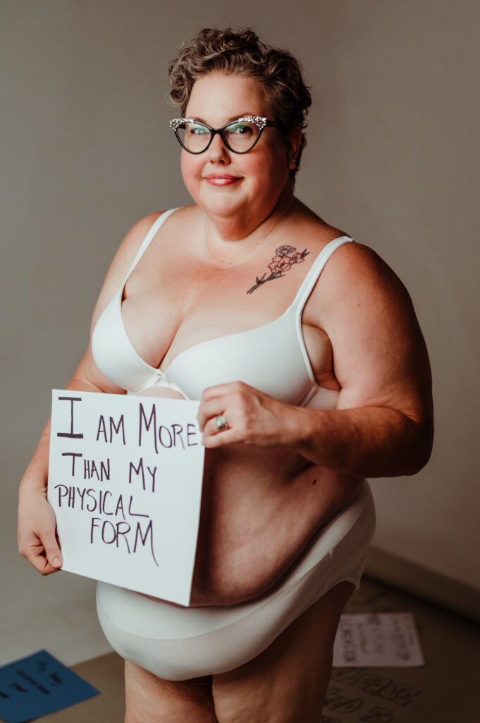 A woman with short curly hair, glasses, and a rose tattoo smiles in white underwear. She holds a sign: I AM MORE THAN MY PHYSICAL FORM. This Nashville boudoir photo radiates an empowering, body-positive message.