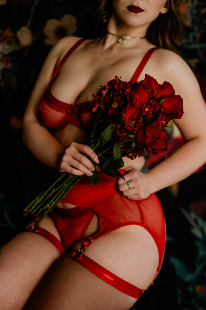A woman in red lingerie holds a bouquet of red flowers against a dark, floral background. Perfect for a Nashville boudoir session, her face is partially out of frame, dark lipstick accenting her turned pose.