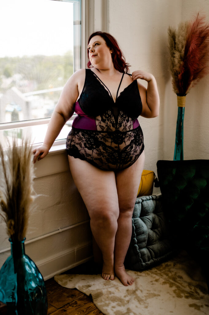 A woman with red hair wearing black lace lingerie stands by a window in a Nashville boudoir, looking outside. She is barefoot, with one hand on her shoulder. The room features plush pillows, a cowhide rug, and tall vases with dried grasses.
