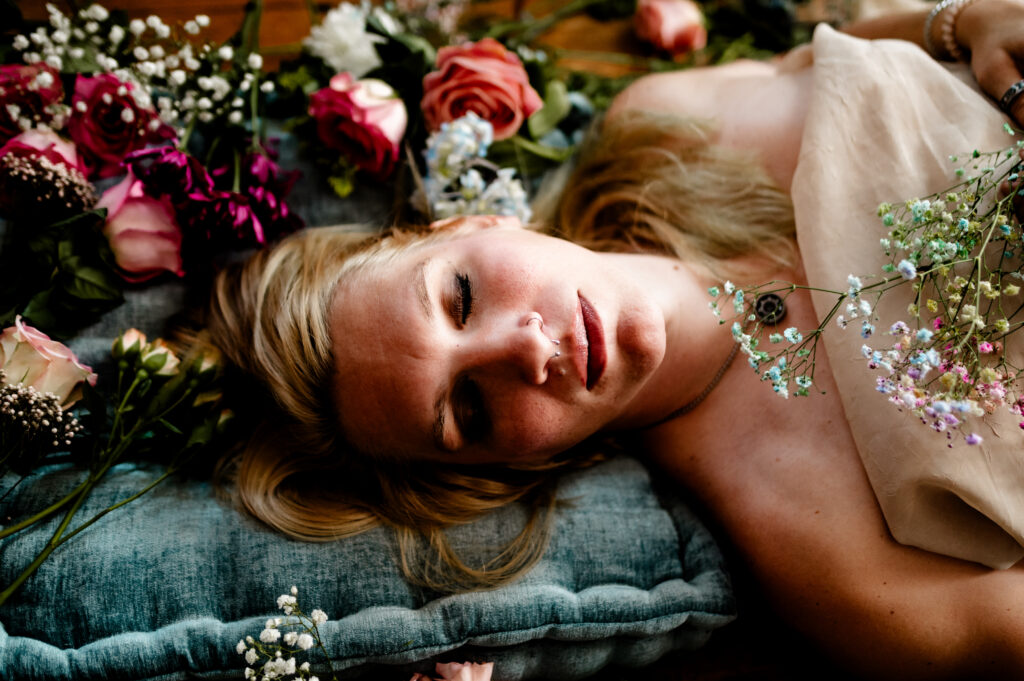 A woman with blonde hair lies on a blue cushion, surrounded by colorful flowers, her eyes closed and peaceful—a serene Nashville boudoir scene.