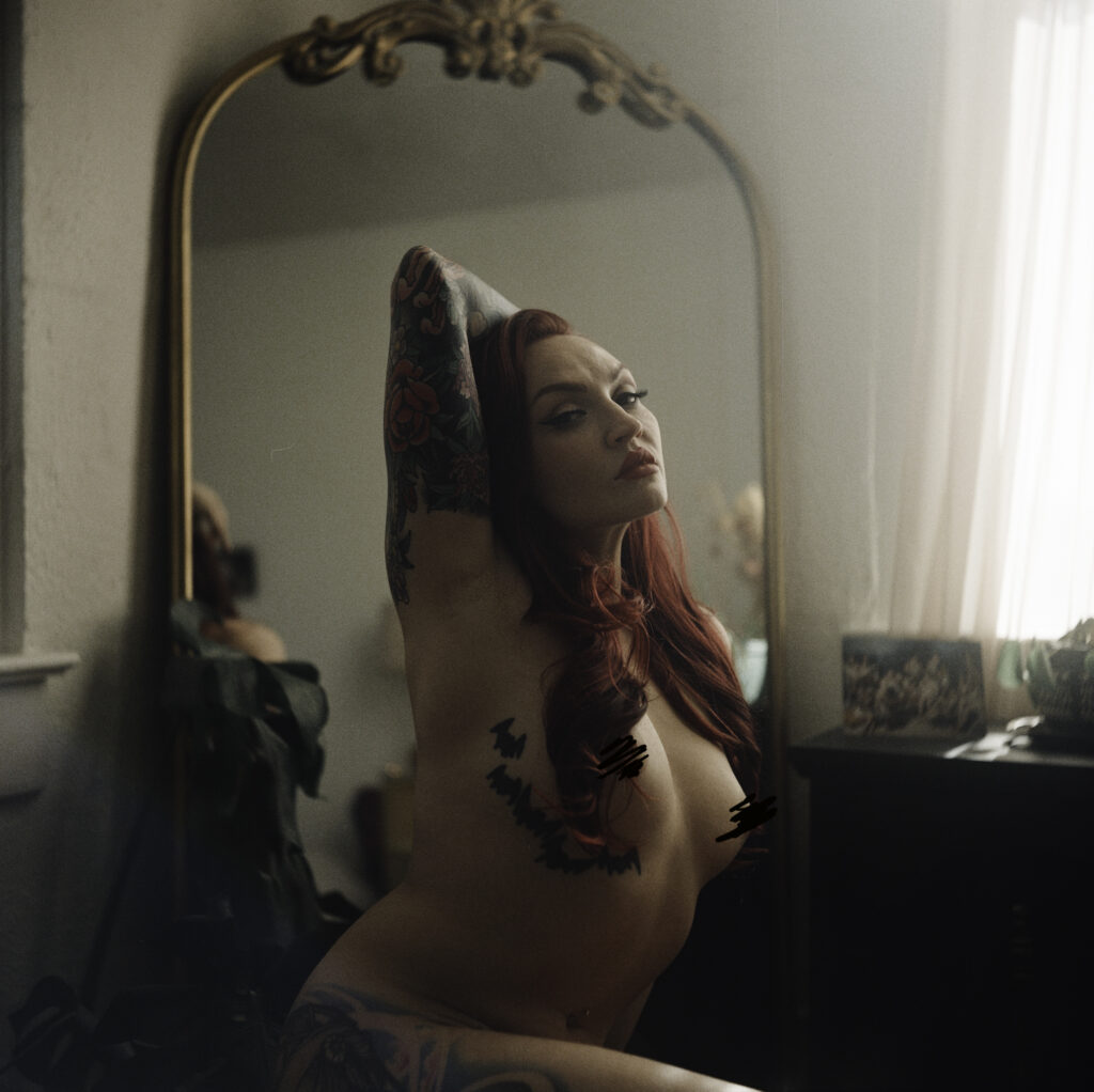 A topless woman with red hair and multiple tattoos poses with one arm raised, sitting in front of an ornate mirror in a softly lit Nashville boudoir.