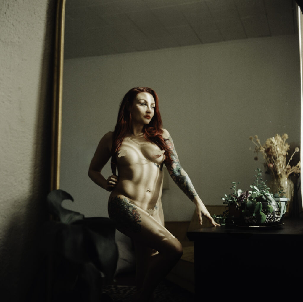 A nude woman with long red hair and colorful tattoos poses confidently by a dresser in front of a mirror, surrounded by plants and dried flowers in a softly lit room—a true expression of Nashville boudoir style.