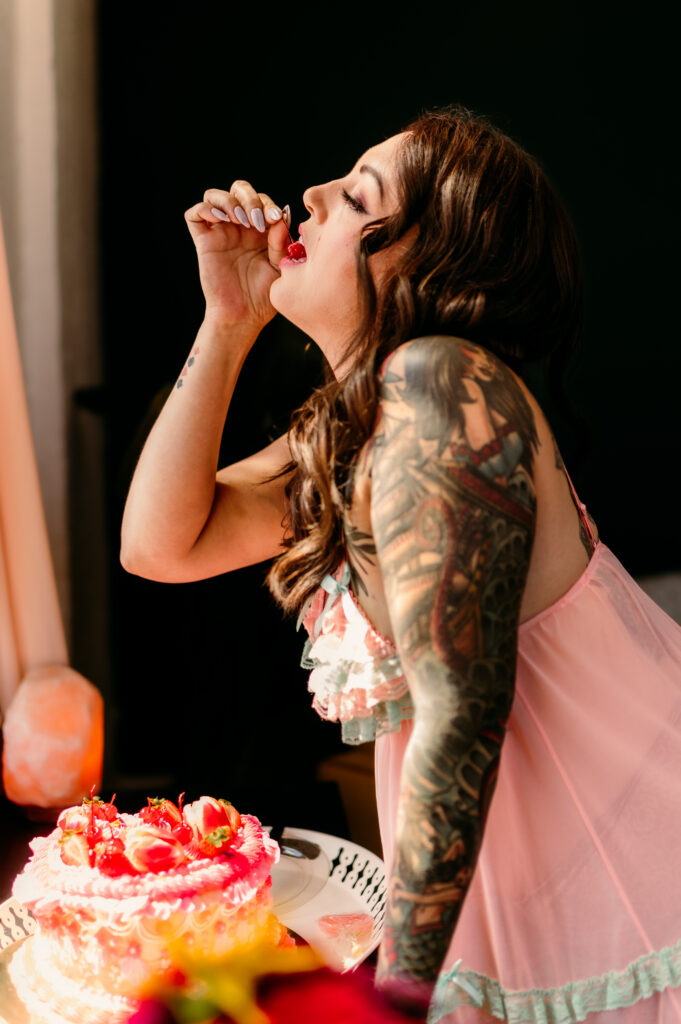 A woman with long brown hair and a tattooed arm, wearing a pink, ruffled dress, is leaning over a decorated cake, about to eat a cherry in a brightly lit room for a playful Nashville boudoir photo shoot.